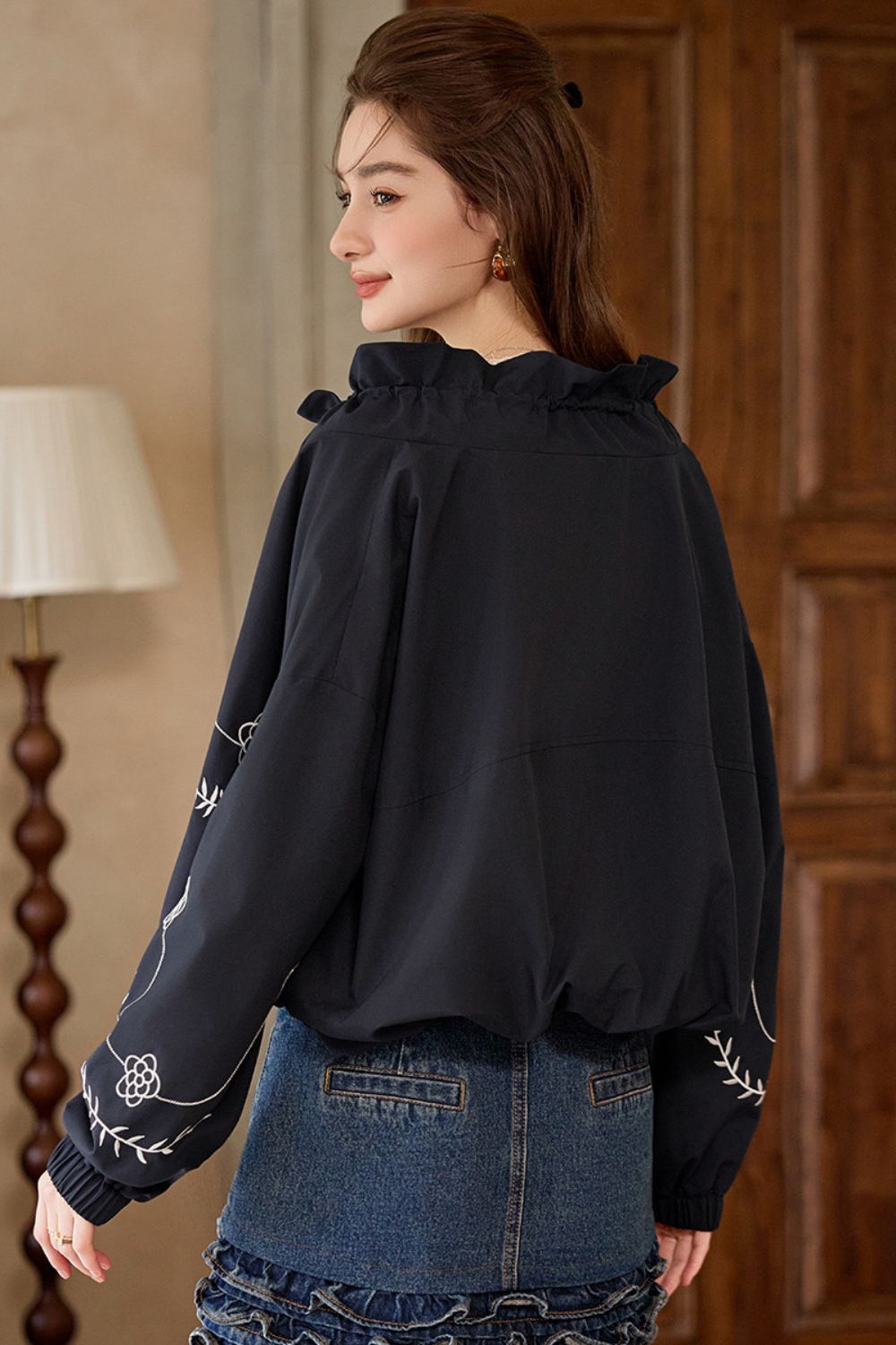 Rear angle view of model wearing black embroidered jacket and denim skirt