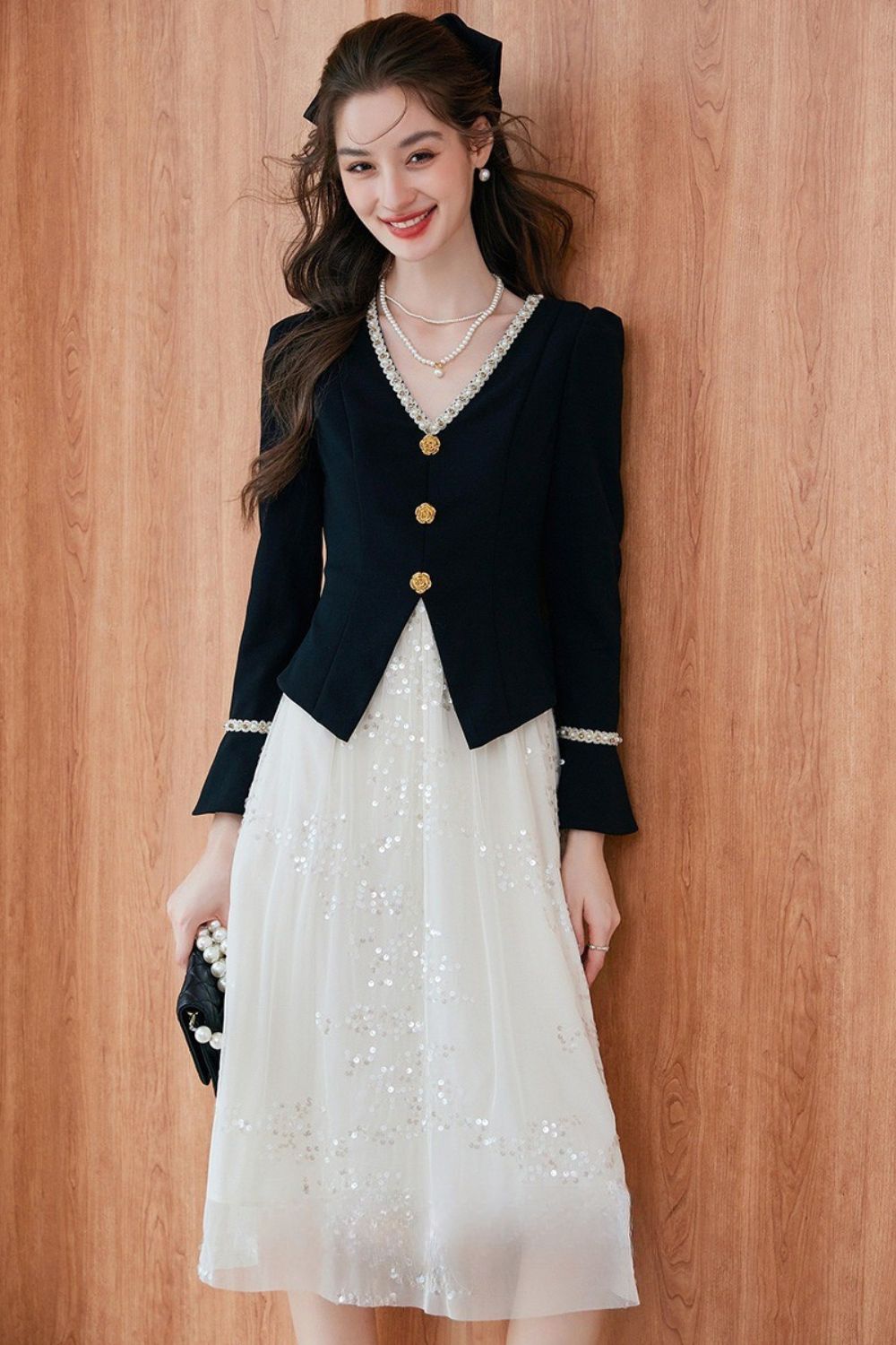 Model wearing a black blazer with gold buttons over a white embroidered midi dress, holding a black handbag