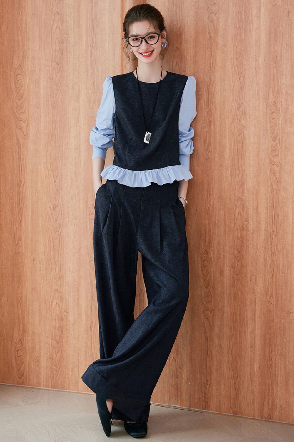 Model wearing a layered outfit with a light blue ruffled blouse under a navy blue sleeveless vest, paired with wide-leg navy trousers