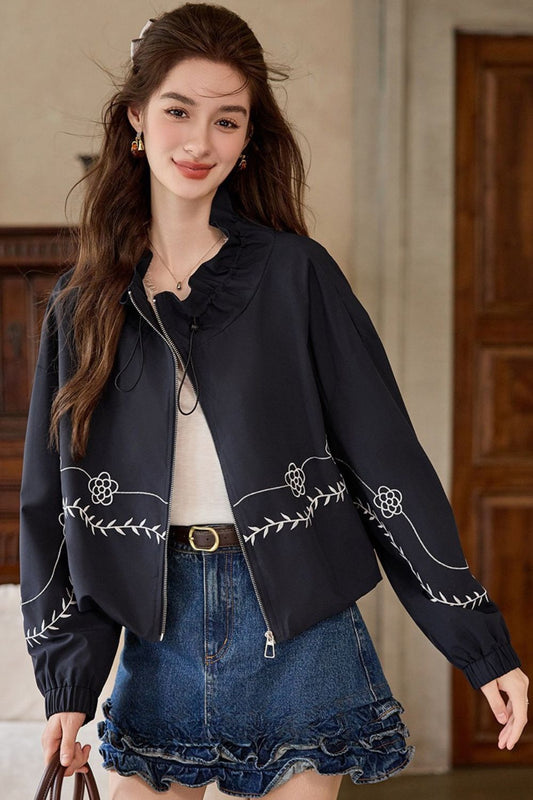 Front view of woman wearing a black embroidered jacket with scalloped trim over a white top and denim skirt