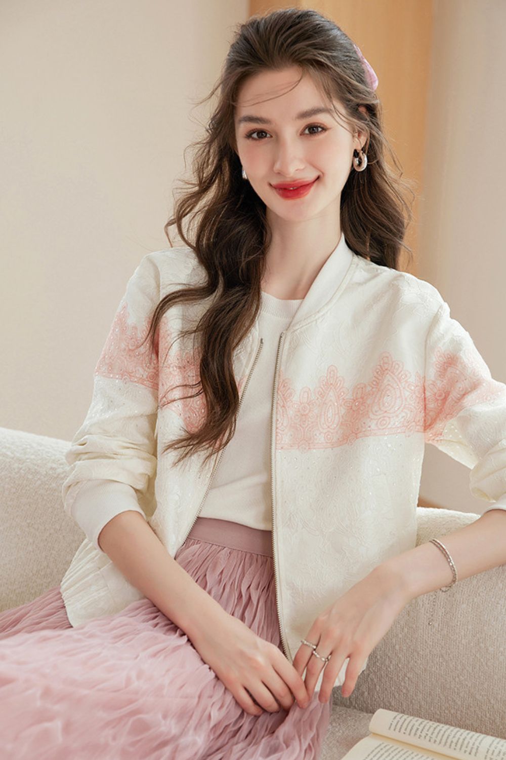 Woman seated in a white cardigan with pink accents and a pink ruffled skirt, with a book on her side