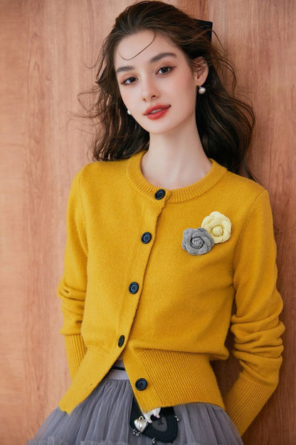 Close-up of model wearing a mustard yellow buttoned cardigan with floral brooch, paired with a gray pleated skirt