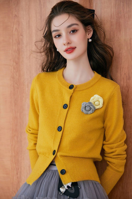 Close-up of model wearing a mustard yellow buttoned cardigan with floral brooch, paired with a gray pleated skirt