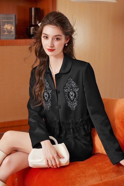 Seated model wearing black embroidered tassel jacket with black skirt