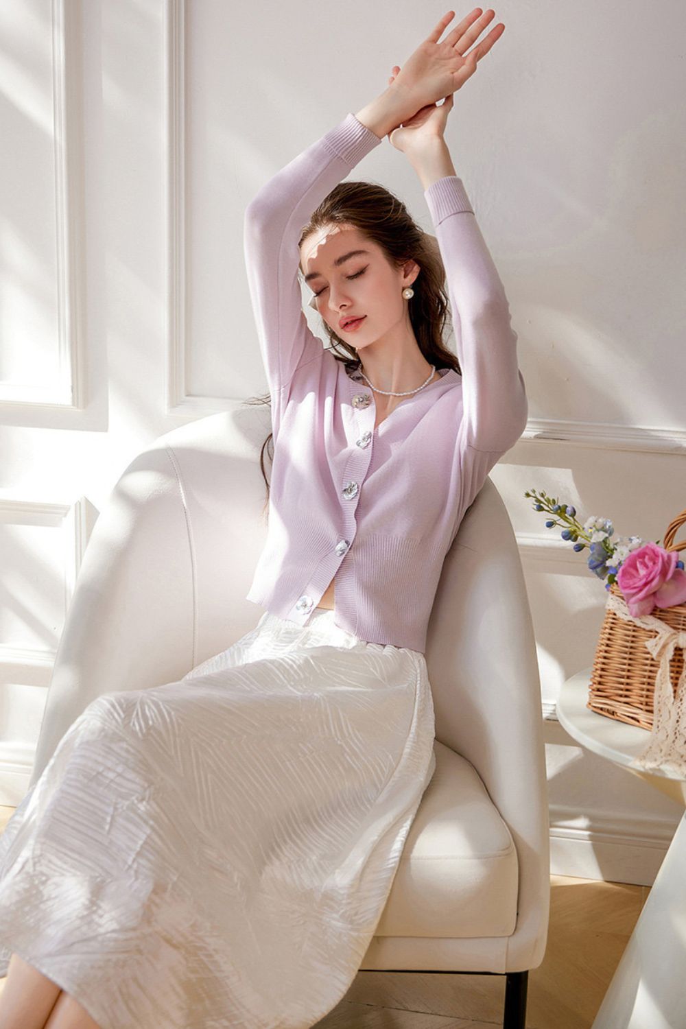 Model stretching while wearing light purple cardigan with pearl buttons and white skirt