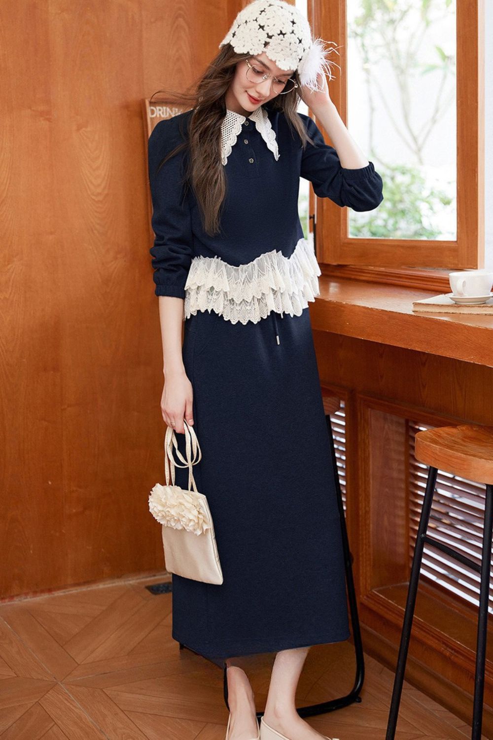 Model leaning on a table wearing navy blue lace-trimmed blouse with navy skirt and crochet hat