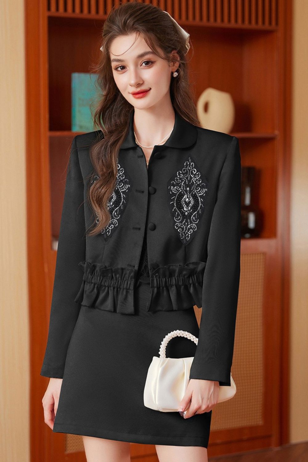 Indoor pose of model in black embroidered jacket with tassels and black skirt