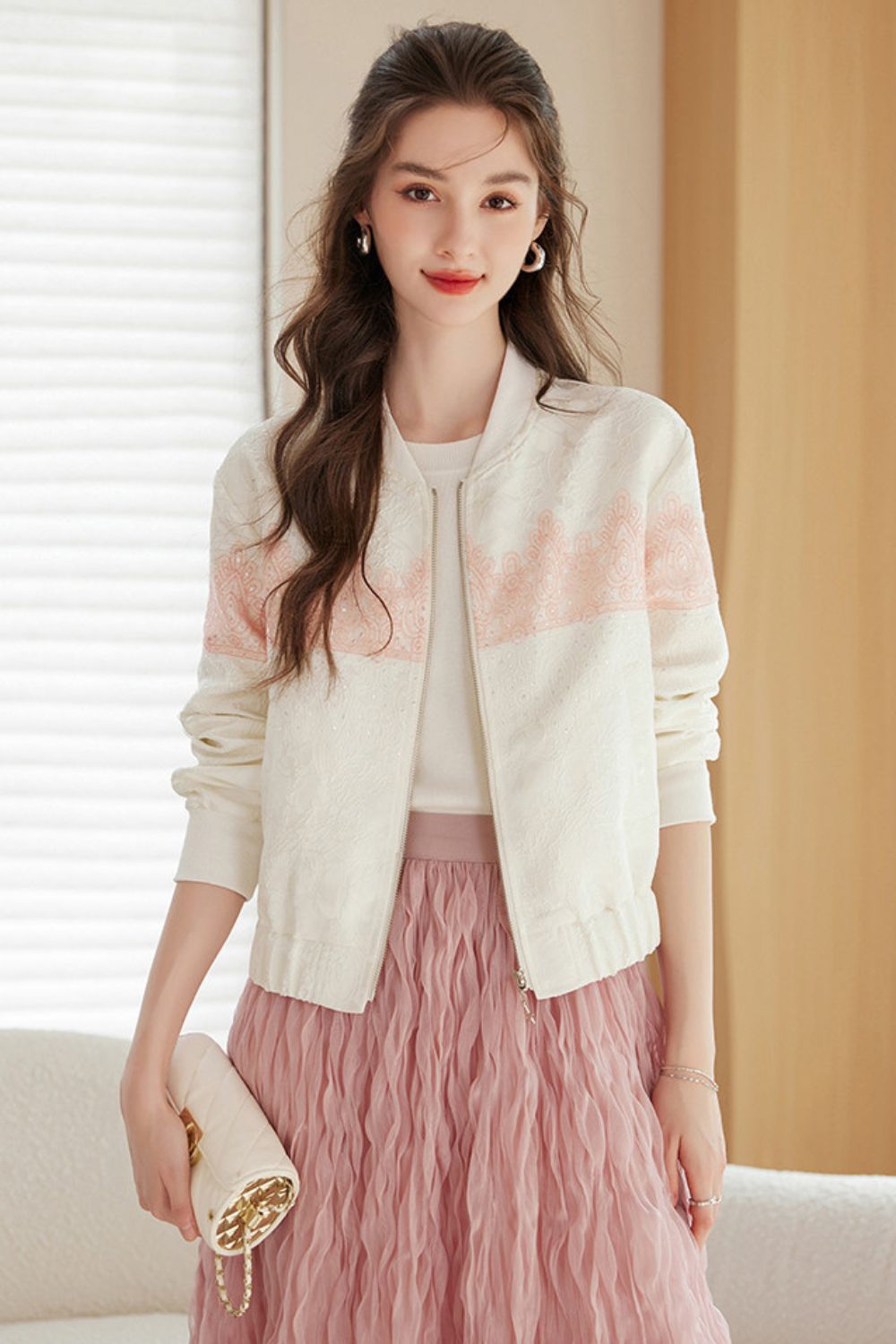 Woman standing and modeling a white cardigan with soft pink accents paired with a pink ruffled skirt