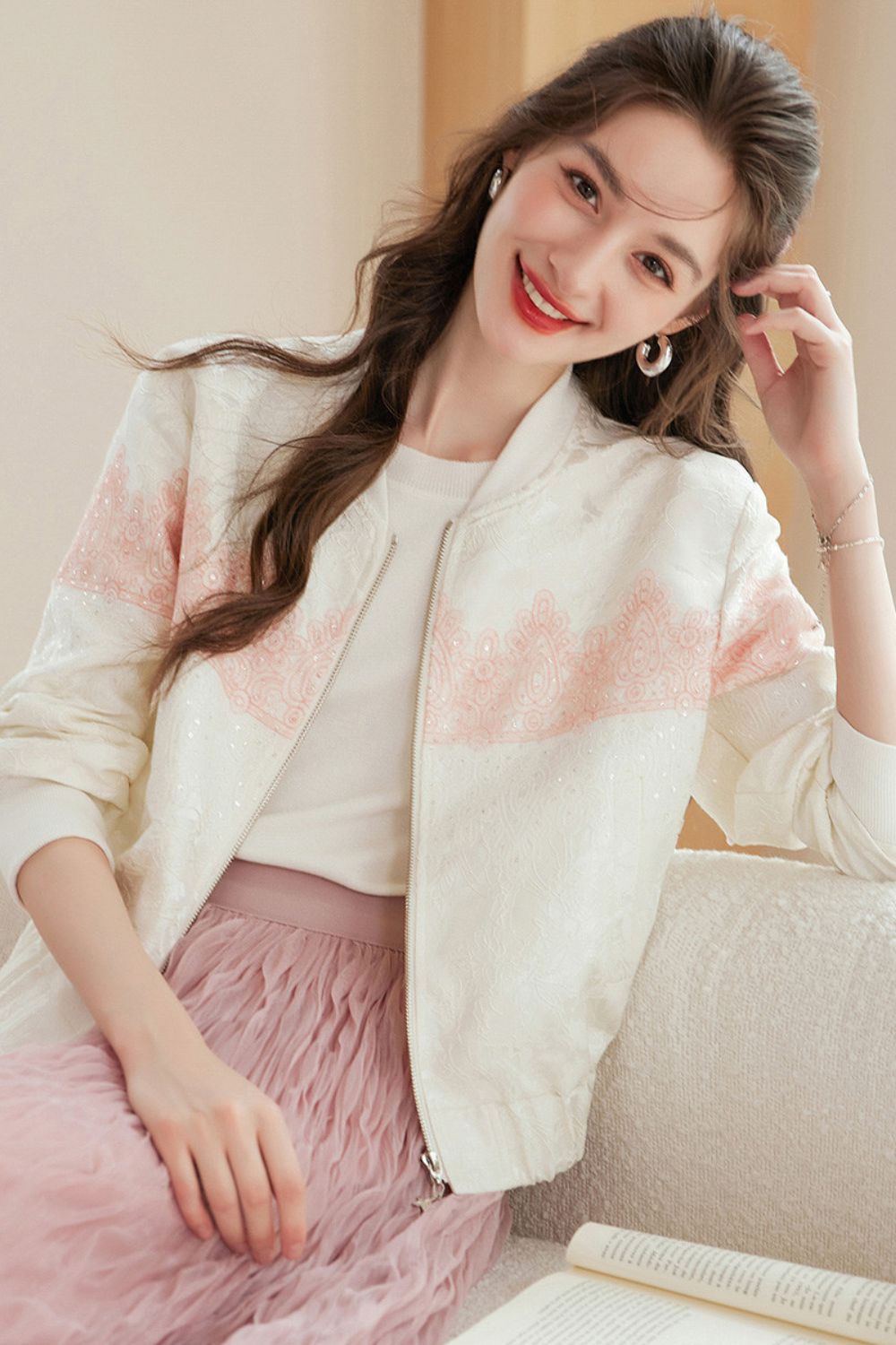 Woman posing on a sofa wearing a white cardigan with pink gradient details and a pink ruffled skirt, hand touching her face