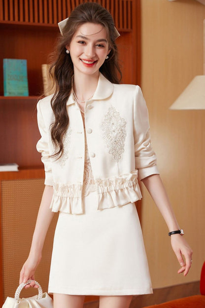 Standing model in cream embroidered tassel jacket and cream skirt holding a handbag