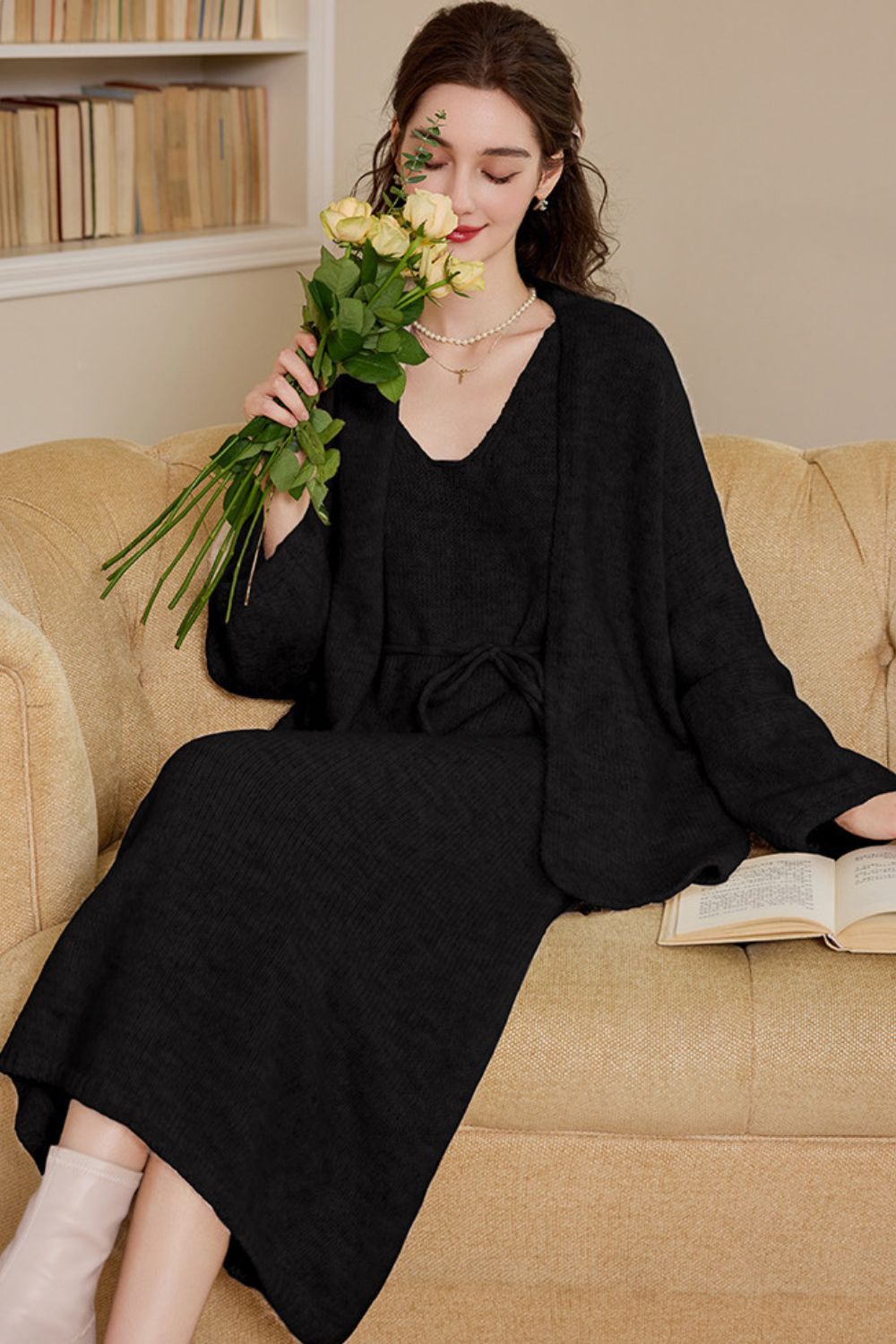 Seated view of black knit cardigan dress on sofa, worn by a woman holding flowers