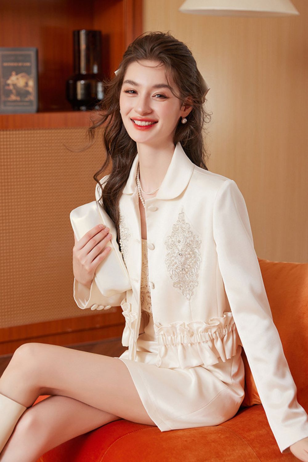 Model seated wearing cream embroidered tassel jacket with cream skirt, smiling indoors